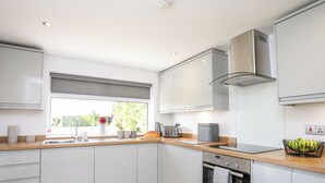A modern kitchen with white cabinets, a wooden countertop, a built-in oven, and a window with a view of greenery outside. - Mynydd Mwyn (Llanfairpwllgwyngyll)