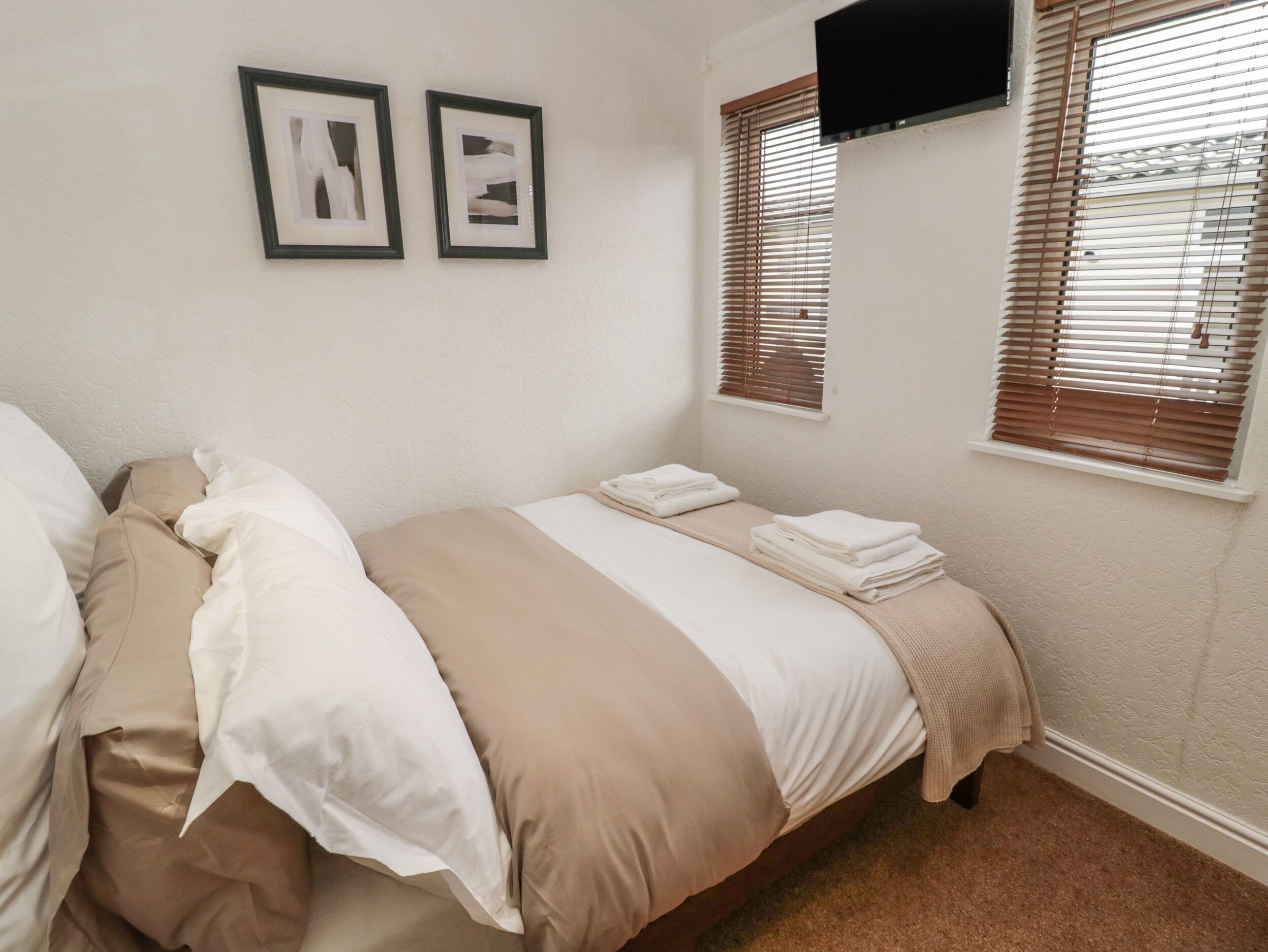 A bedroom with a bed, two framed pictures on the wall, a TV mounted above the bed, and a window with blinds.