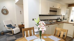 A dining area with a round wooden table, six chairs, and a vase with flowers. There is a kitchen in the background with white cabinets and a black backsplash.