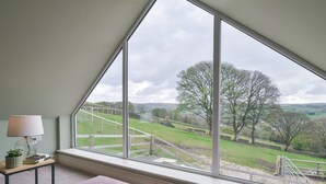Een kamer met een groot raam dat uitzicht geeft op een landelijk landschap, een kleine tafel met een lamp, en twee paarse poefs.