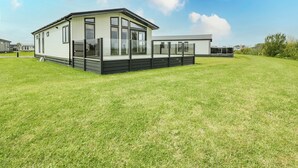 A modern mobile home with a large deck, surrounded by a grassy field.