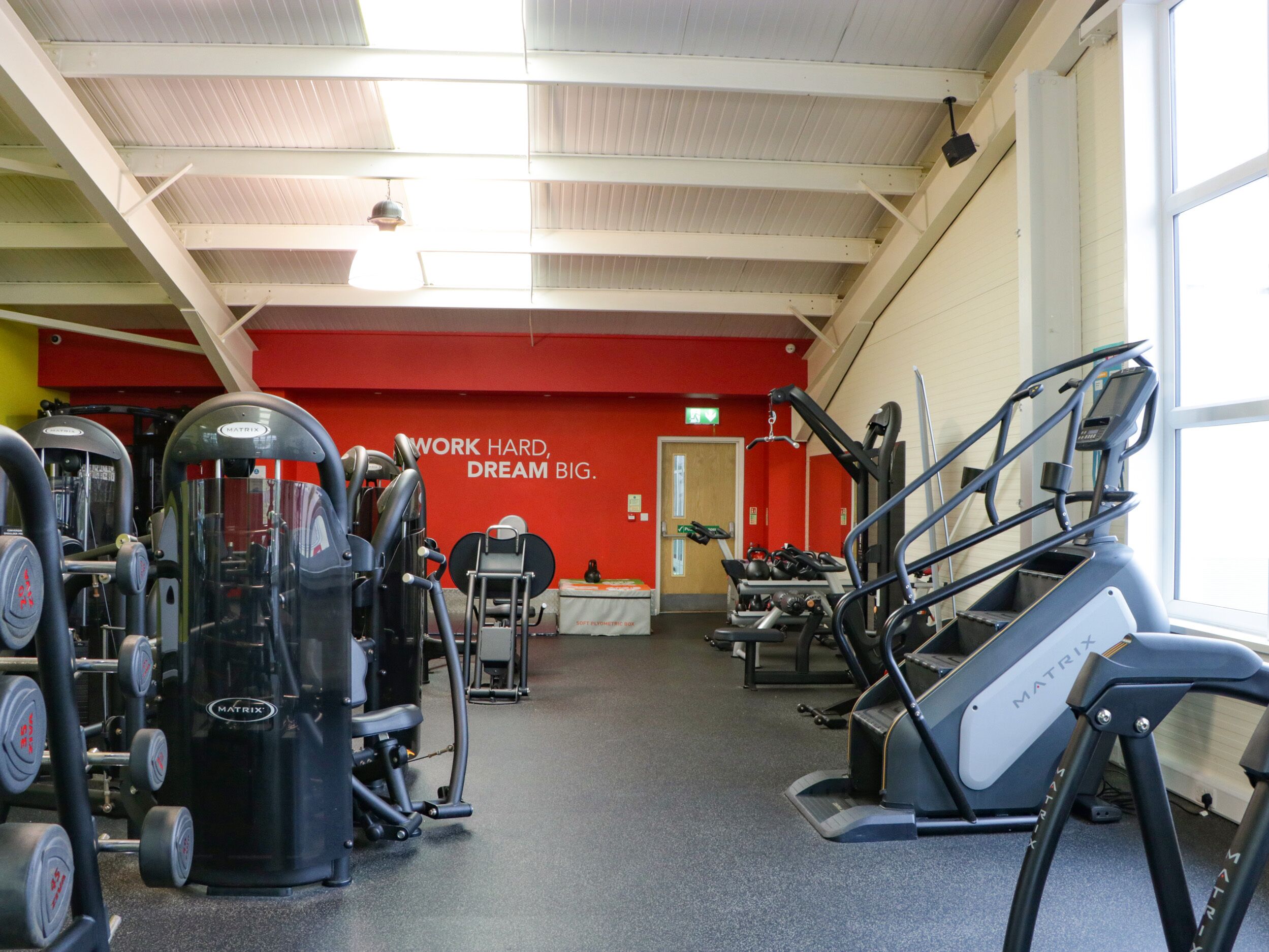 Une salle de sport équipée de divers appareils d’exercice, avec un mur rouge décoré de messages motivants et une fenêtre laissant entrer la lumière naturelle.