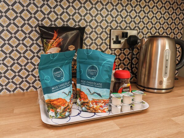Una encimera de cocina con dos bolsas de Chocolate con Leche Guppy's, un tarro de Nescafé Original y una tetera de acero inoxidable.