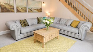 A living room with a grey sofa, a wooden coffee table, and a yellow patterned rug. There are three paintings on the wall and a staircase in the background.