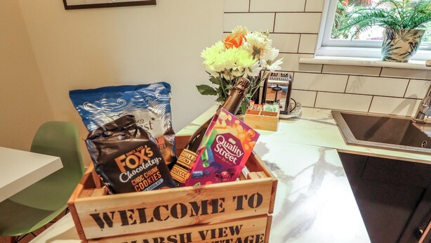 A wooden welcome sign in a kitchen area with a variety of items including a bag of chocolate cookies, a bottle, and a bouquet of flowers.
