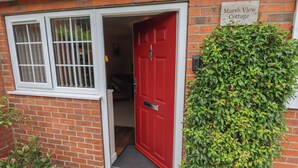 A red door with a sign reading "Marsh View Cottage," a brick wall, a window with white frames, and a green hedge.