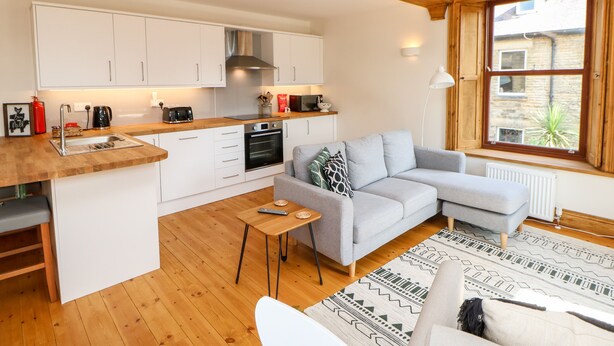 Ein kompakter Wohnraum mit Küchenzeile, einem grauen Sofa, einem kleinen Tisch und einem gemusterten Teppich.