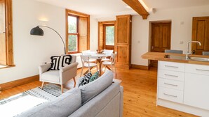 Eine moderne Küche mit Holzschränken, einer weißen Arbeitsplatte und einem Spülbecken. Es gibt einen Essbereich mit Tisch und Stühlen sowie ein Wohnzimmer mit Sofa und Lampe.