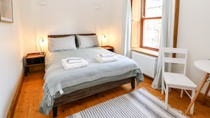 Ein Schlafzimmer mit einem hölzernen Bett, zwei Nachttischen jeweils mit einer Lampe, einem Fenster mit Vorhängen, einem weißen Stuhl und einem kleinen runden Tisch.