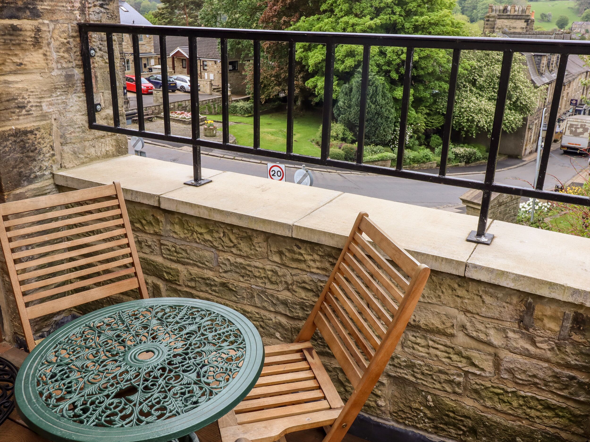 A balcony with two wooden chairs and a table, overlooking a street with parked cars and greenery.