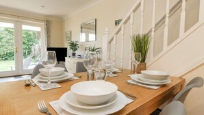 Una mesa de comedor dispuesta con platos, tazones y cubiertos blancos, junto a una escalera y vistas a zonas verdes al exterior.
