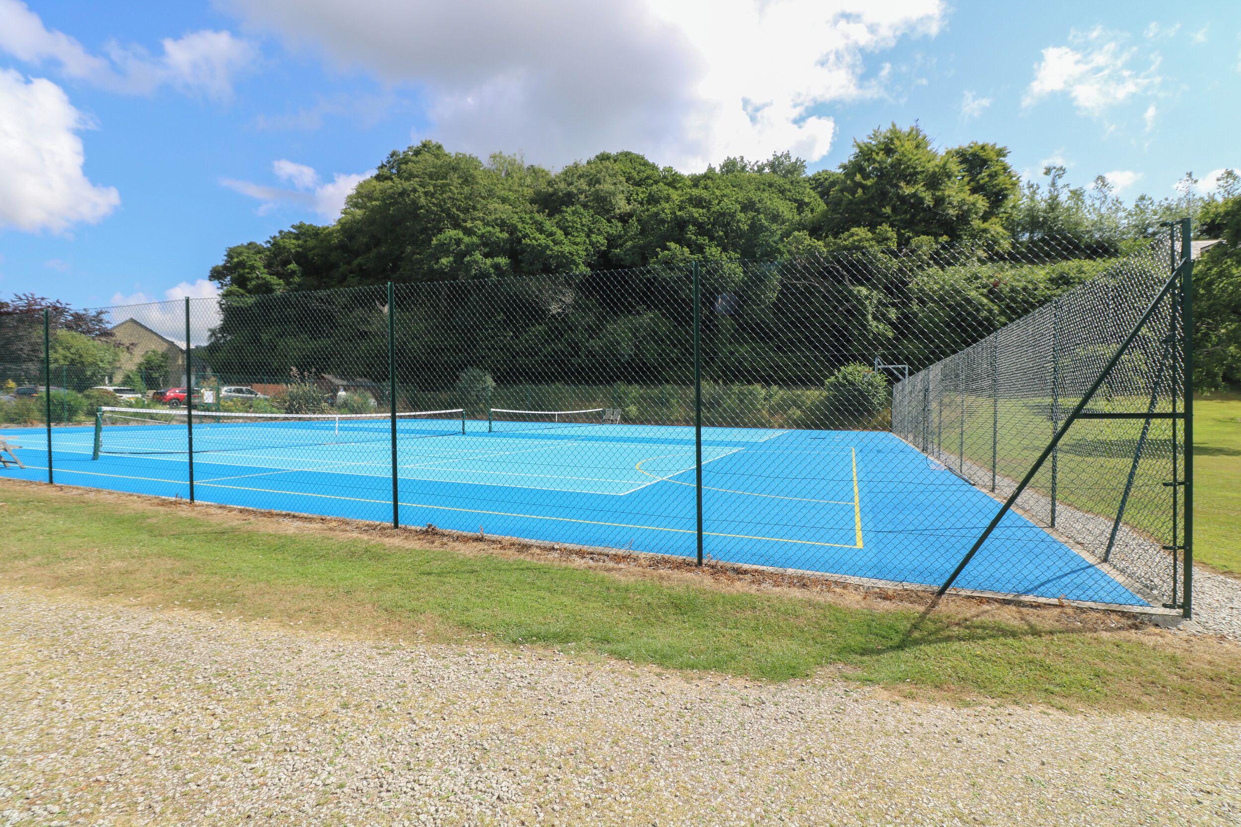An outdoor tennis court with a chain-link fence, surrounded by trees and a grassy area.