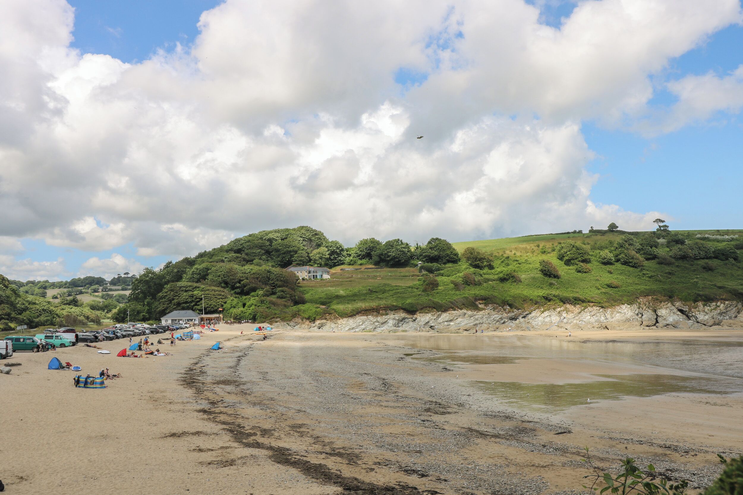 A beach with tents and cars, a house on a hill, and a partly cloudy sky.