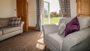 A living room with two sofas, one with a plaid cushion and another with a purple cushion, a wooden door, and a view of a garden through a glass door. - Hillcrest (Retford)