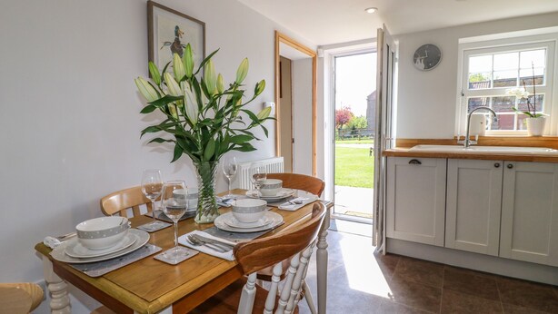 A dining area with a wooden table set for four, a vase with flowers, and a view of the garden through an open door. - Hillcrest (Retford)