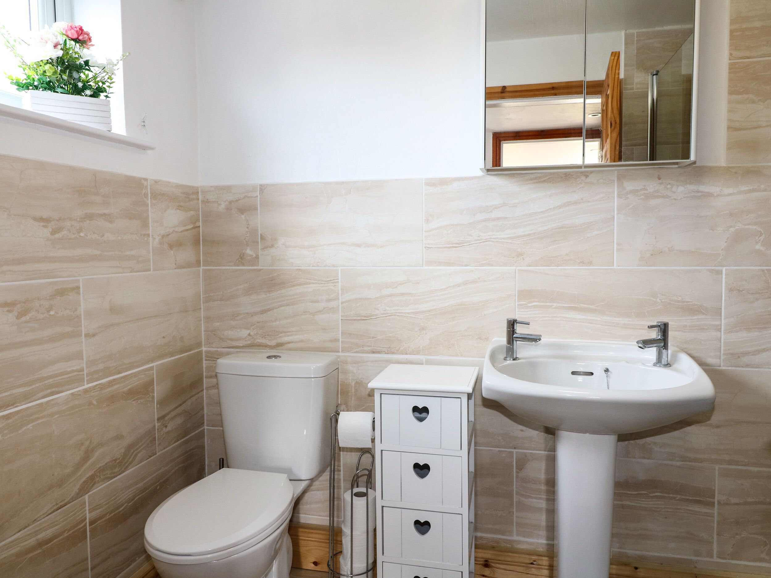 Une salle de bain avec des carreaux de couleur beige, des toilettes blanches, un lavabo blanc et un miroir au-dessus du lavabo.