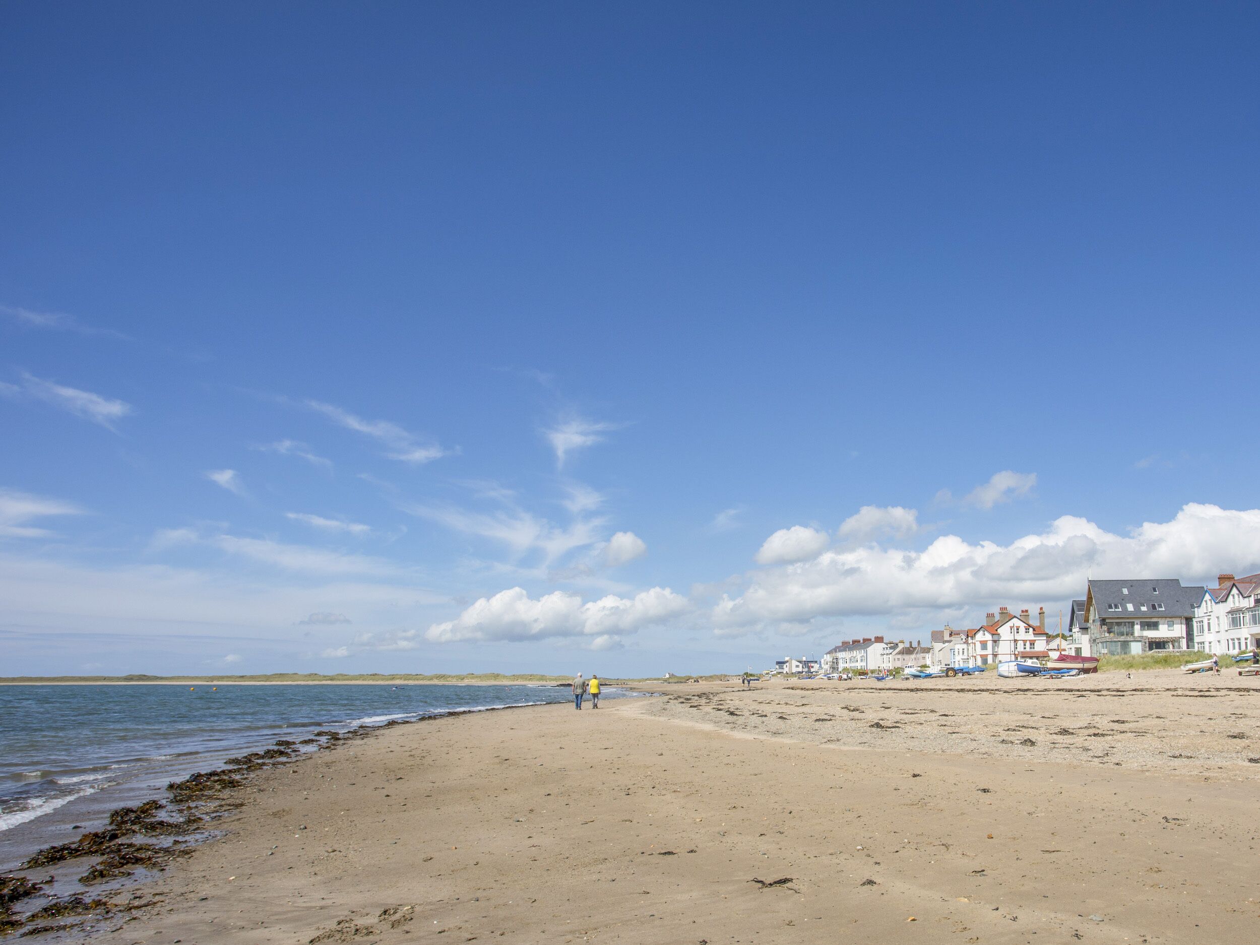 Een zandstrand met blauwe lucht, enkele mensen die wandelen en huizen op de achtergrond.