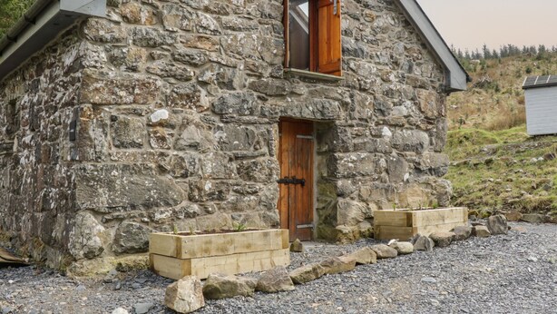 Una casa in pietra con una porta e una finestra in legno, circondata da un vialetto sterrato e piccoli fiorieri in legno.