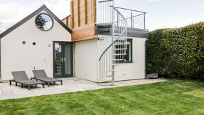 A modern house with a small patio, two lounge chairs, a wooden deck, and a metal staircase. - Vale Lodge (Pewsey)
