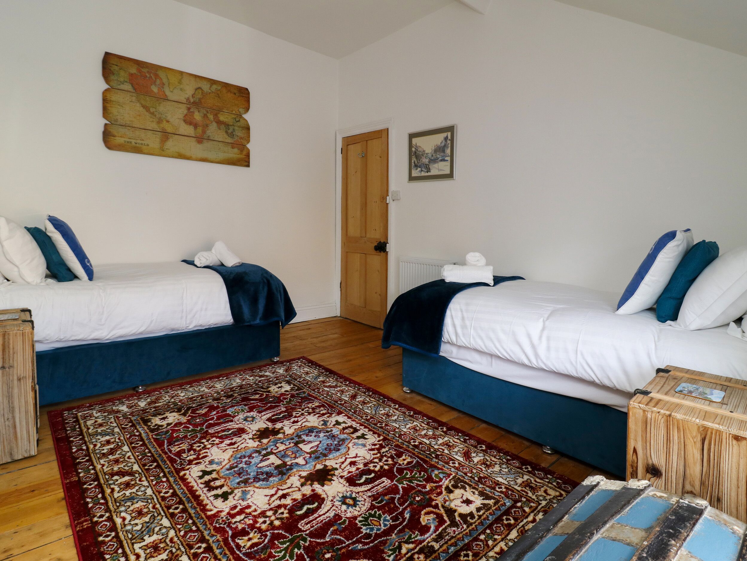A room with two beds, a wooden floor, a red patterned rug, and a painting on the wall.