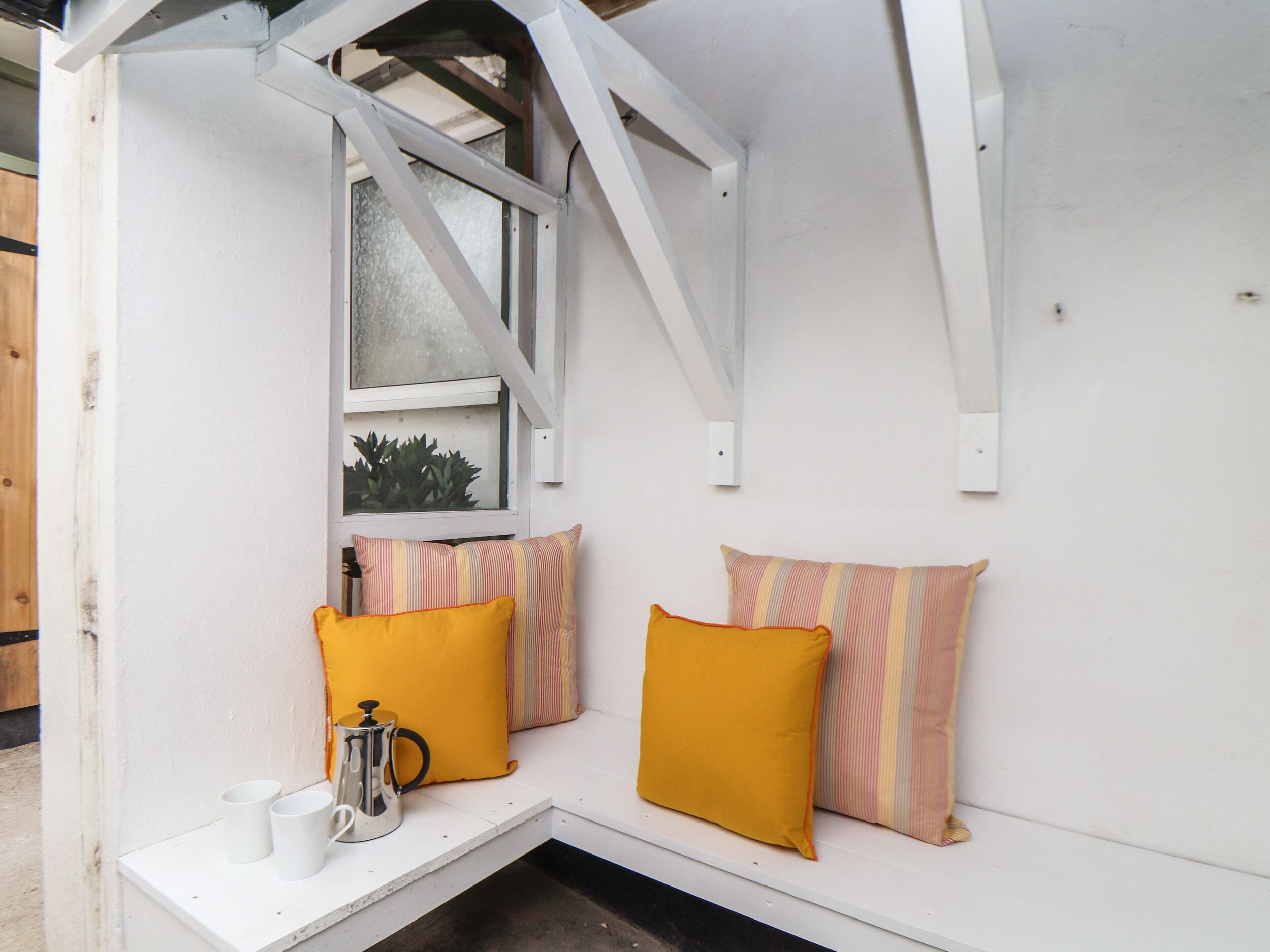 Un rincón de descanso blanco y cerrado con una ventana, dos almohadas amarillas y una taza blanca en una estantería.