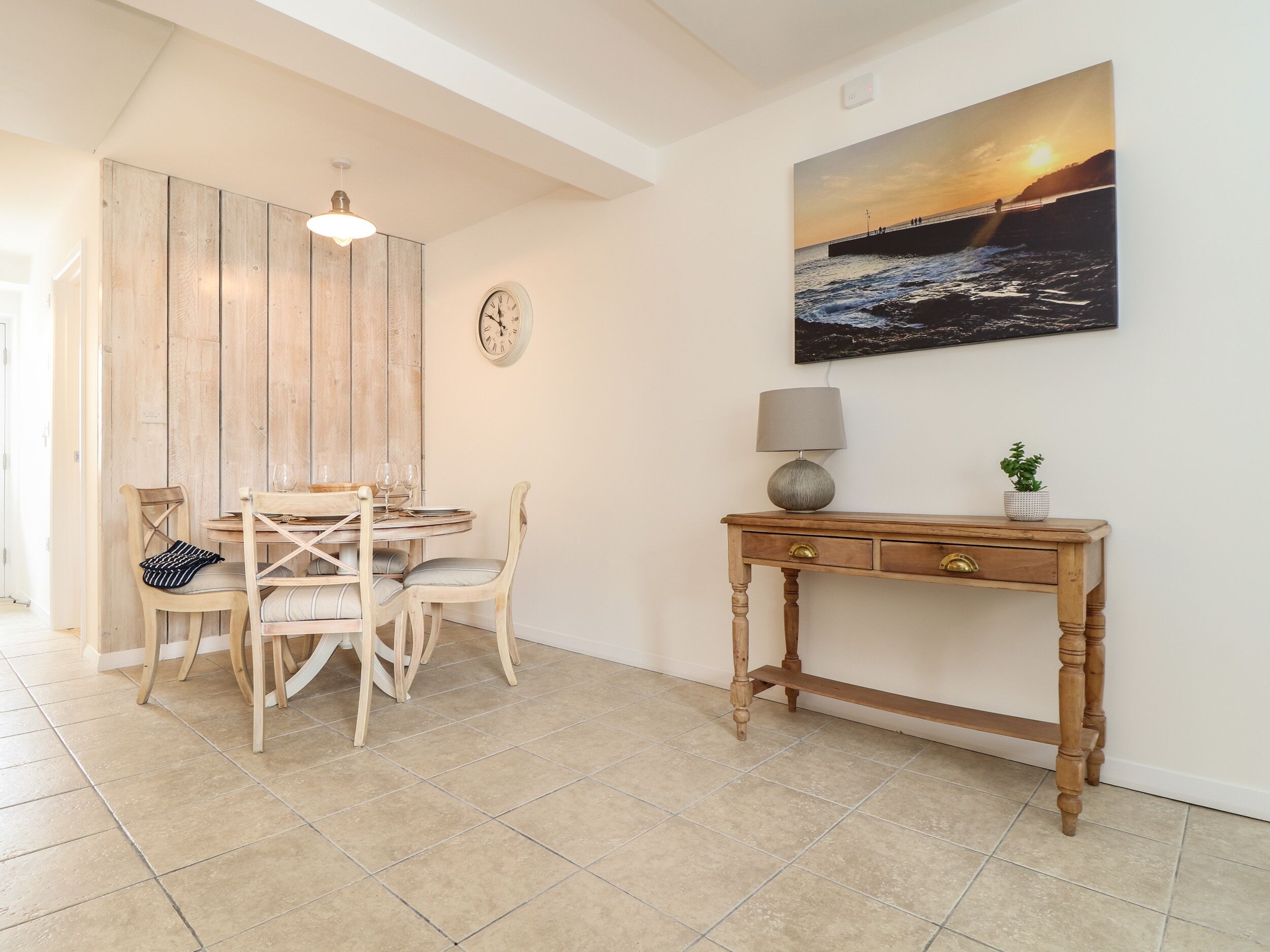 Uma área de refeições com mesa e cadeiras de madeira, um relógio de parede, uma pintura de pôr do sol e uma pequena mesa com abajur e planta.