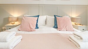 Een keurig opgemaakt bed met roze en blauwe kussens, een wit hoofdbord en een muur met drie ingelijste foto's.