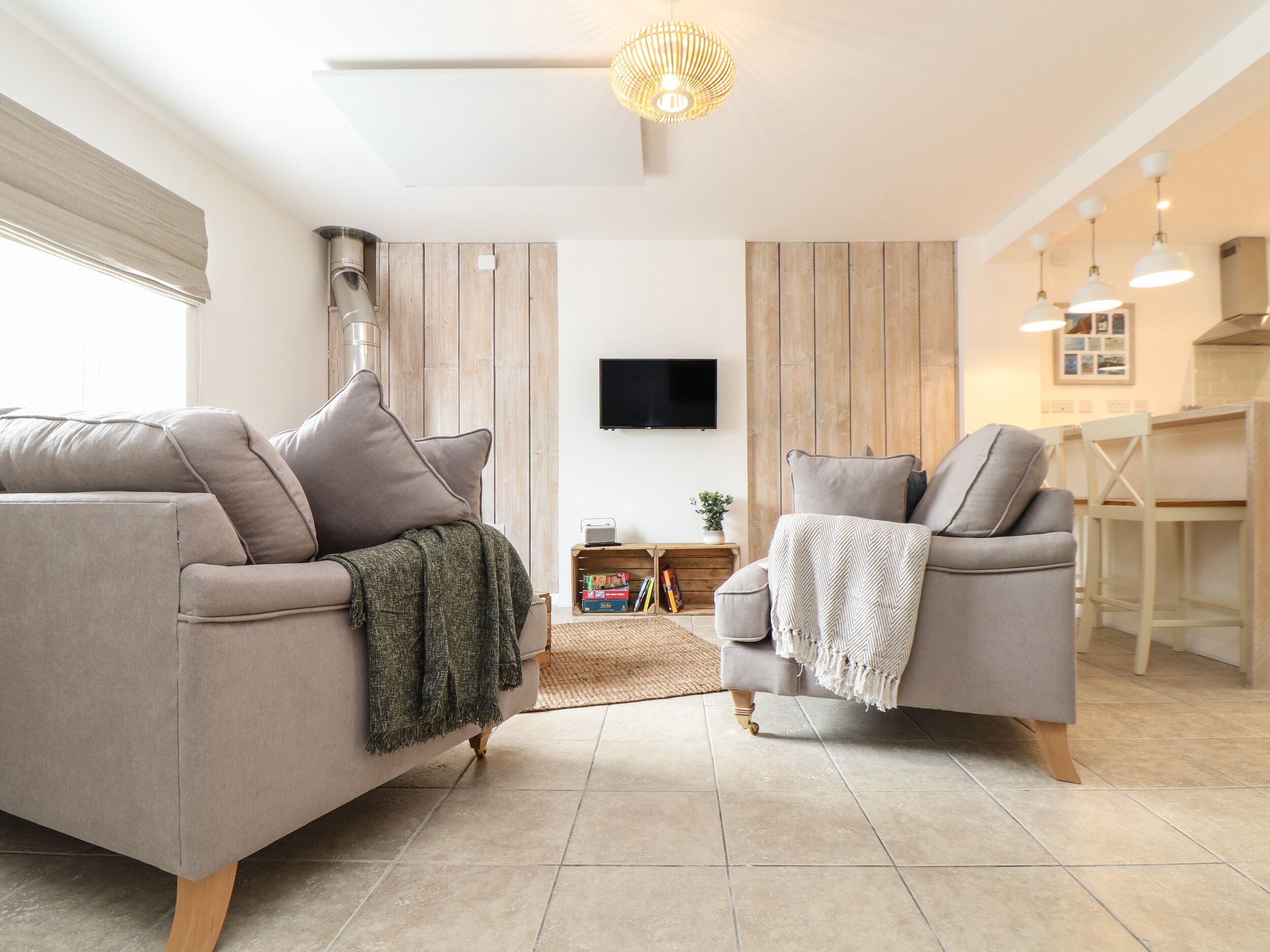 A modern living room with two sofas, a flat-screen TV, wooden panel walls, and a kitchen area with pendant lights.