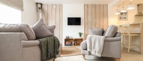 A modern living room with two sofas, a flat-screen TV, wooden panel walls, and a kitchen area with pendant lights.
