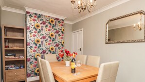 A dining room with a wooden table, beige chairs, a floral wallpaper, a bookshelf, and a mirror on the wall. - Poppy Cottage (Deal)