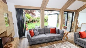 Un salon moderne avec un canapé gris, des coussins rouges et gris, une table basse en bois et une grande fenêtre donnant sur un jardin et une maison.