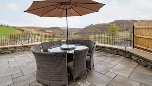 Une terrasse extérieure avec une table ronde et des chaises, un grand parasol et une vue panoramique sur des collines et une route.