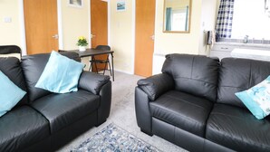 A room with two black leather sofas, a small table with a vase of flowers, and a kitchen area with a sink and window. - 20 Siesta Mar (Norwich)