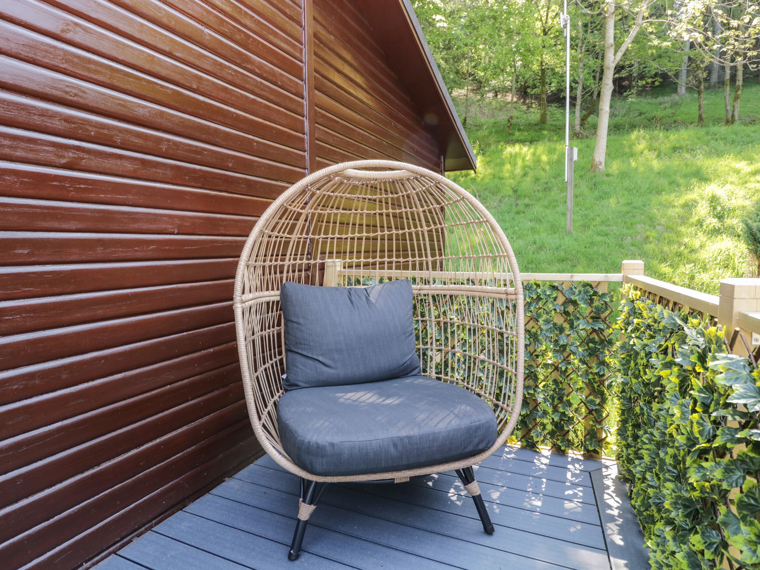 A wicker chair with a cushion on a wooden deck, adjacent to a wooden building with a sloped roof.