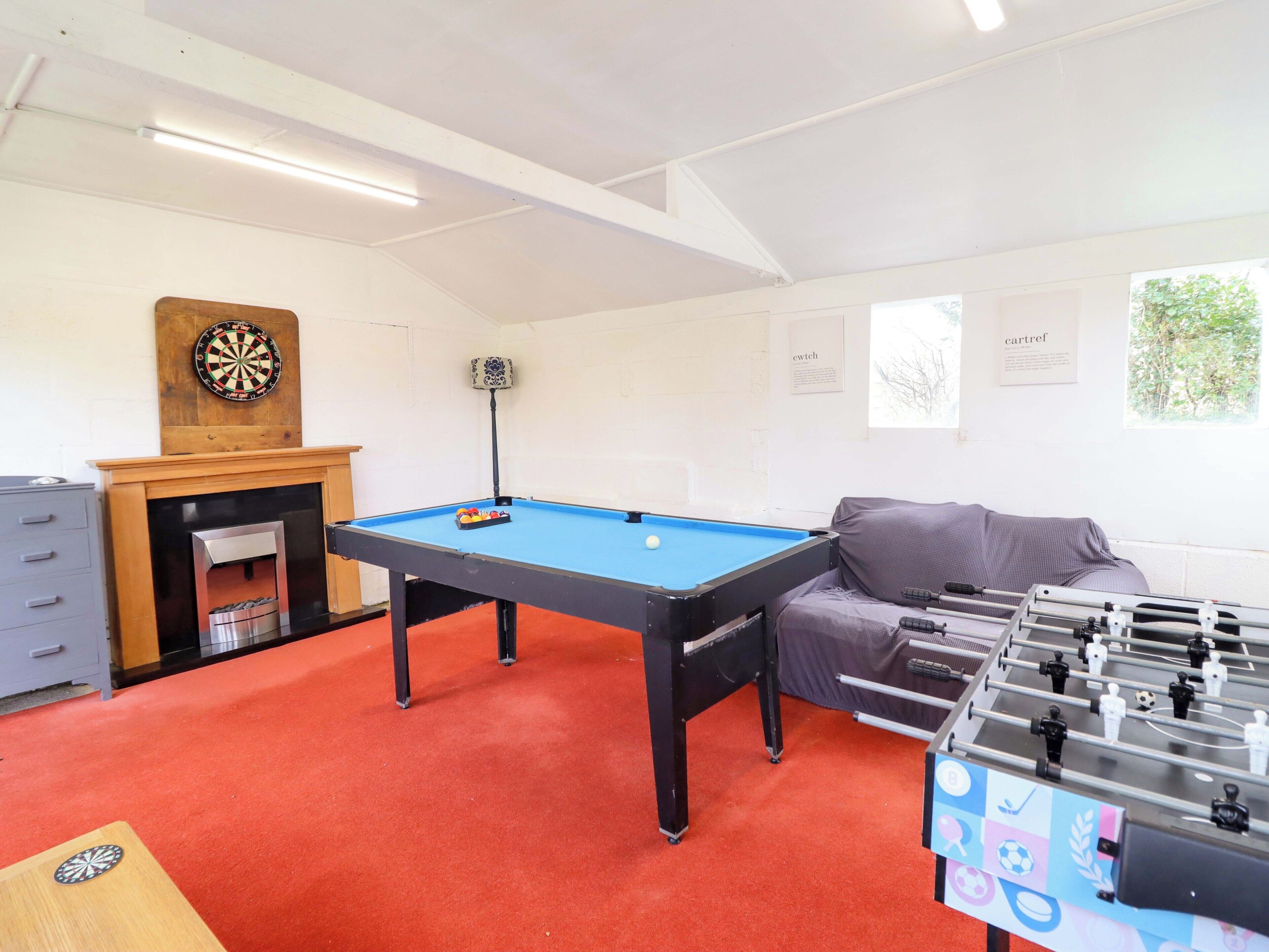A room with a pool table, a dartboard, a foosball table, and a fireplace.