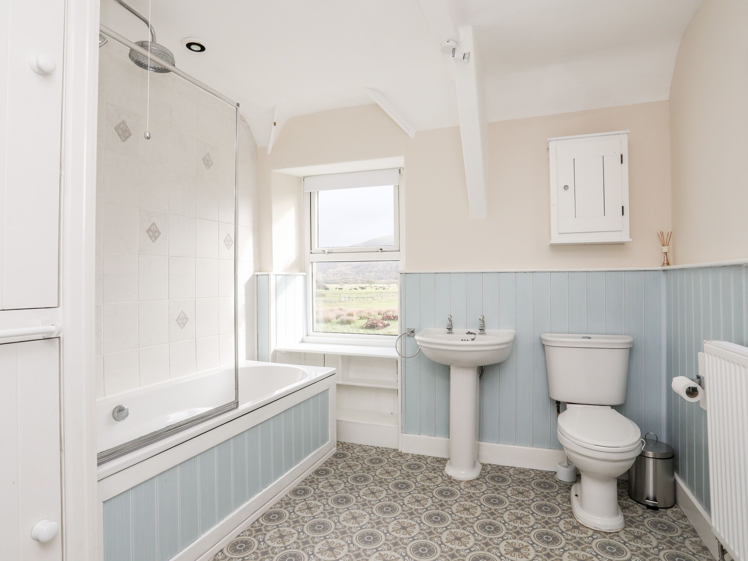 A bathroom with a shower, sink, toilet, and a window with a view of the countryside.