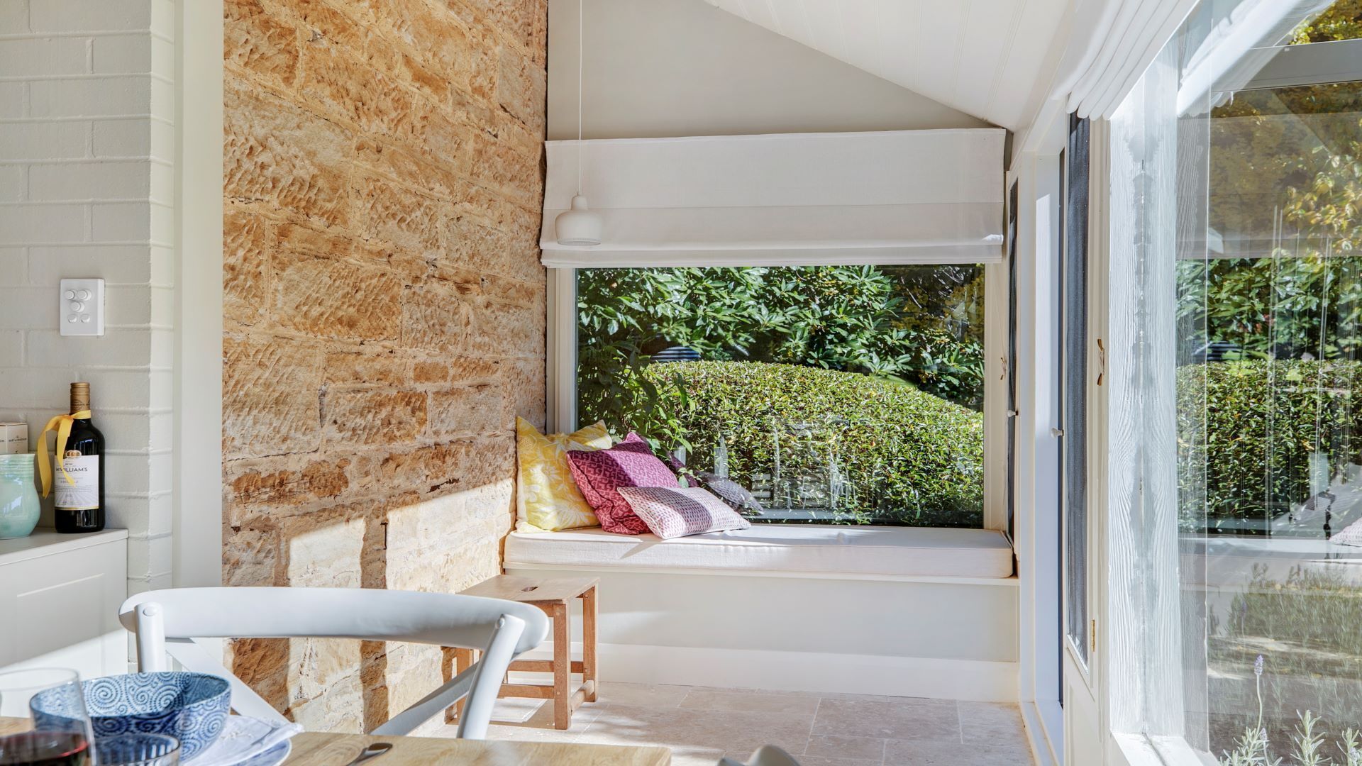 Una habitación acogedora con pared de piedra, ventana con vistas a la vegetación, una silla blanca y una mesa con una botella y vasos.