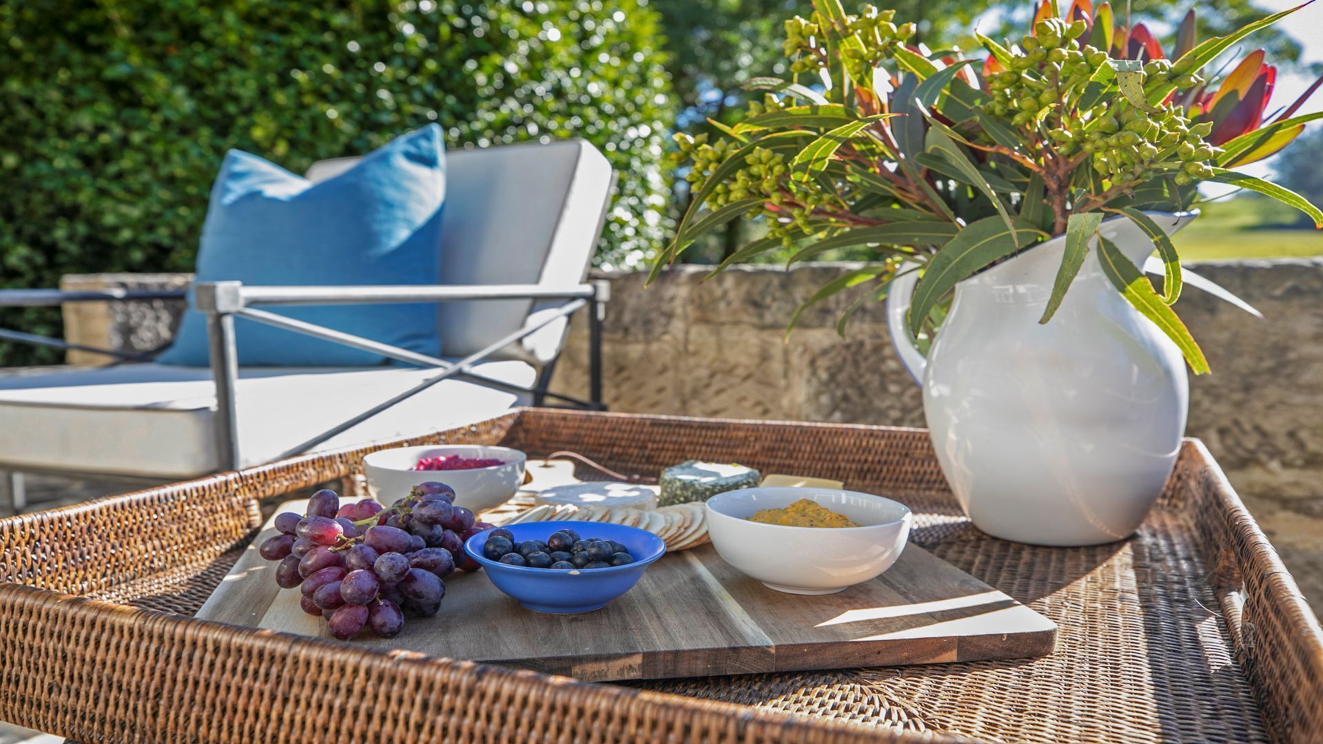 A wicker tray with a variety of food items including grapes, blueberries, and cheese, placed on an outdoor patio with a chair and a vase of flowers in the background.