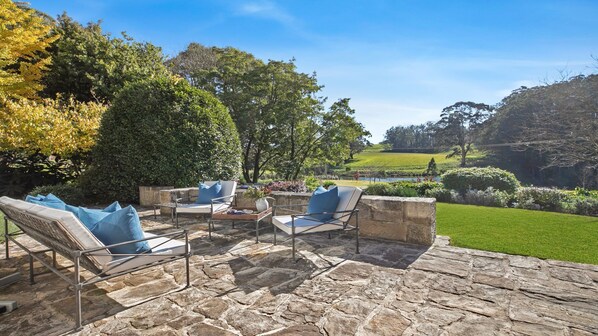 Une terrasse extérieure avec un sol en pierre, deux fauteuils de jardin dotés de coussins bleus, et une vue sur un jardin luxuriant et des collines verdoyantes en arrière-plan.