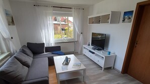 En moderne stue med en grå sofa, et sofabord, et fjernsyn på et hvidt skab og et vindue med gardiner.