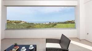 En balkon med havudsigt og grønne planter, en fletstol og et bord med en bog og kopper.