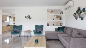 En moderne stue med en grå sofa, to turkise lænestole, et træ sofabord og et lille rundt bord. En trappe med hvid gelænder og en reol med dekorative genstande ses i baggrunden.