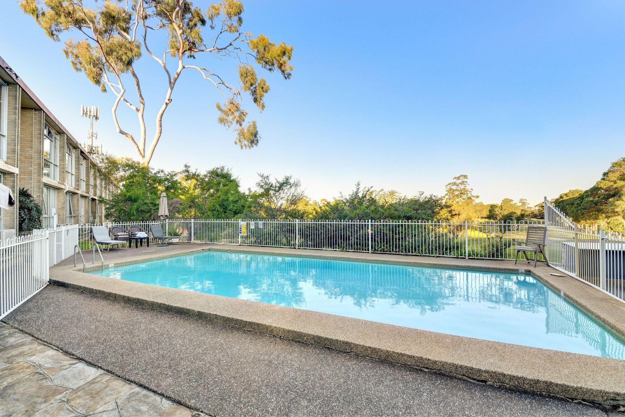 Una piscina rectangular con valla blanca, rodeada por una zona pavimentada con sillas de descanso y un edificio con techo de tejas.