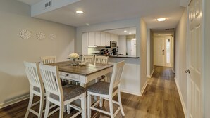Un comedor con mesa de madera y sillas blancas, una cocina al fondo y un pasillo que lleva a una puerta.