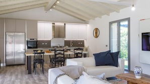 A modern kitchen with a built-in oven and a stainless steel refrigerator. There is a dining area with a table and chairs, and a sofa with a blue pillow.
