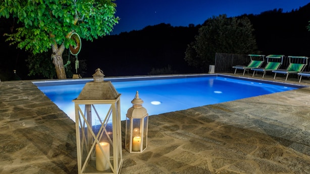 An outdoor swimming pool with stone flooring, two lanterns, and a tree with green leaves.