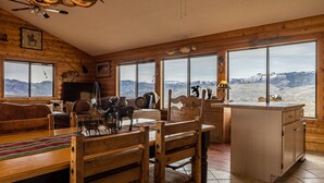 Interior rústico de una cabaña con mobiliario de madera, chimenea y ventanas amplias con vistas a las montañas.