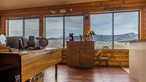 Interior rústico de una cabaña con paredes de madera, ventanas amplias que ofrecen vistas a la montaña, un escritorio de madera y un armario.