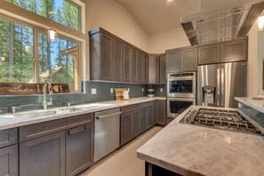 A modern kitchen with dark cabinets, stainless steel appliances, and a large window overlooking a forest.