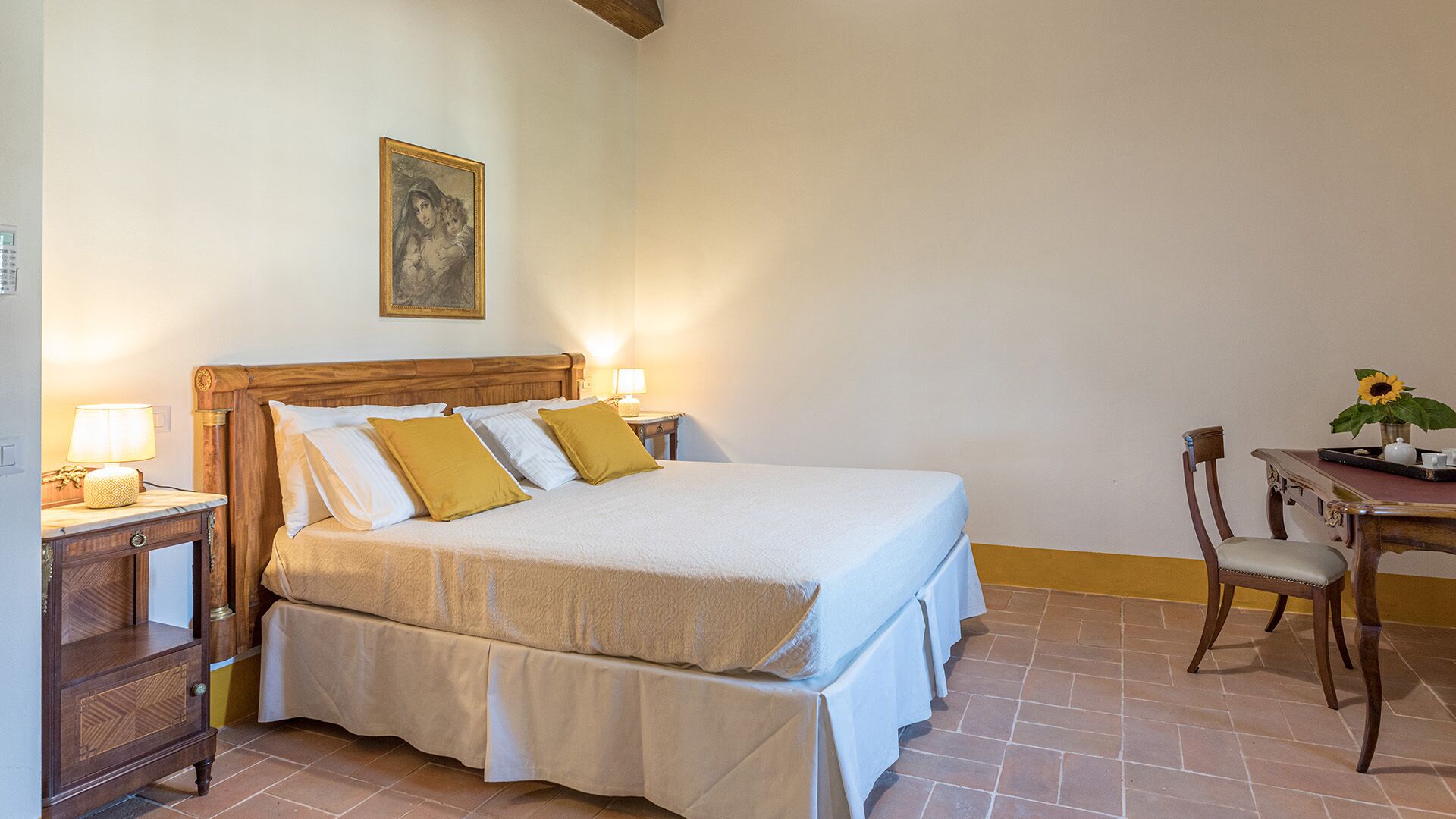 Una cama bien hecha con sábanas blancas y almohadones amarillos, una mesita de noche de madera con una lámpara, un cuadro en la pared y una pequeña mesa con un jarrón de girasoles.